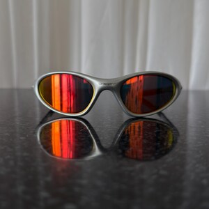 Puede incluir: Gafas de sol con montura plateada y lentes espejadas rojas y naranjas vibrantes. Las gafas de sol se reflejan en una superficie oscura y brillante. El logotipo de Oakley es visible en la montura.