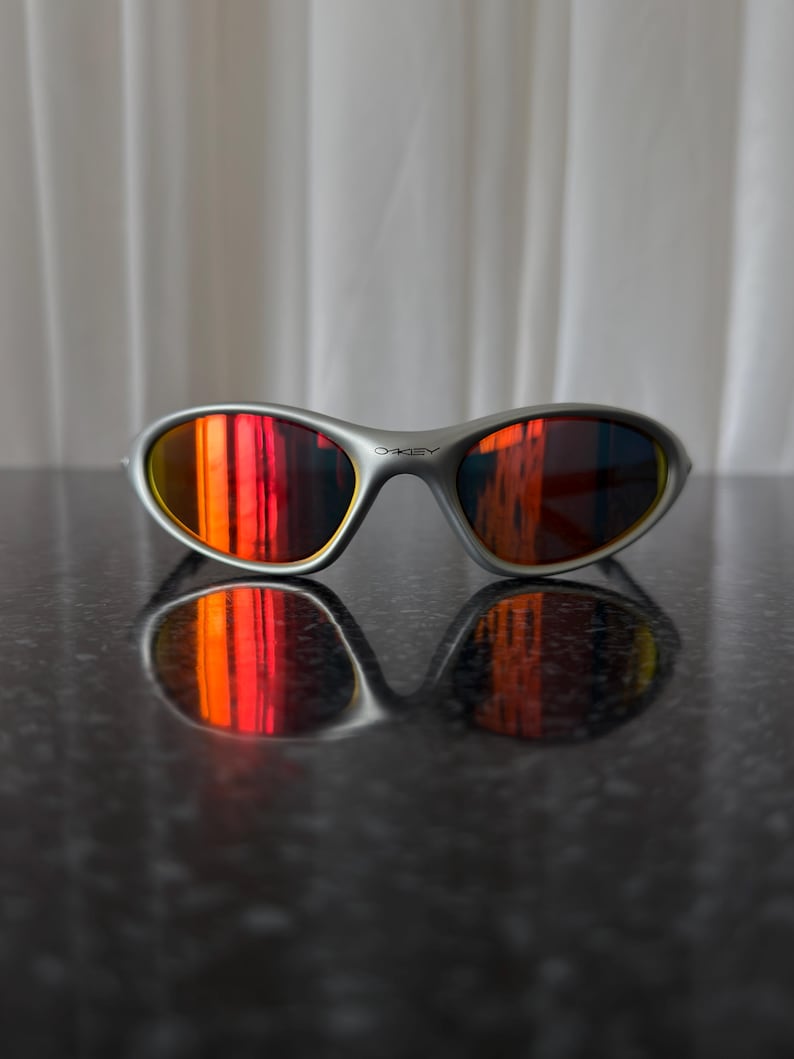 Puede incluir: Gafas de sol con montura plateada y lentes espejadas rojas y naranjas vibrantes. Las gafas de sol se reflejan en una superficie oscura y brillante. El logotipo de Oakley es visible en la montura.