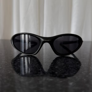 K&ouml;nnte beinhalten: Schwarze, oval geformte Sonnenbrille mit dunklen Gl&auml;sern. Die Sonnenbrille liegt auf einer reflektierenden, dunklen Oberfl&auml;che, mit einem wei&szlig;en Vorhang im Hintergrund. Das Design ist modern und elegant.