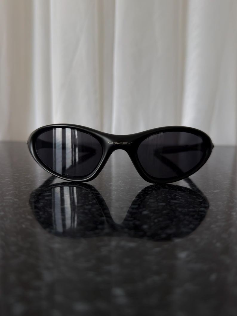 K&ouml;nnte beinhalten: Schwarze, oval geformte Sonnenbrille mit dunklen Gl&auml;sern. Die Sonnenbrille liegt auf einer reflektierenden, dunklen Oberfl&auml;che, mit einem wei&szlig;en Vorhang im Hintergrund. Das Design ist modern und elegant.