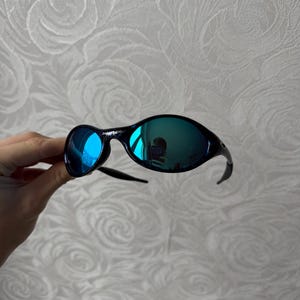 K&ouml;nnte beinhalten: Schwarze Sonnenbrille mit blau verspiegelten Gl&auml;sern. Die Sonnenbrille hat einen geschwungenen Rahmen und ein schlankes Design. Die Gl&auml;ser reflektieren die Umgebung und erzeugen einen lebendigen, farbenfrohen Effekt.