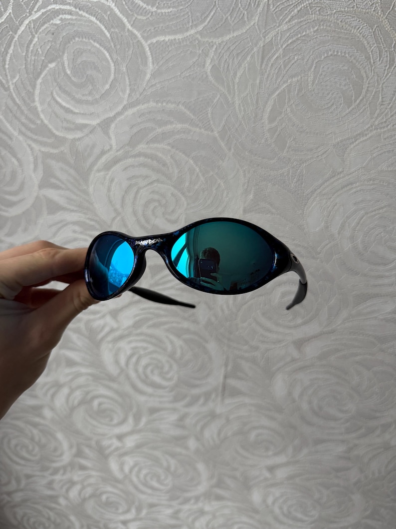 K&ouml;nnte beinhalten: Schwarze Sonnenbrille mit blau verspiegelten Gl&auml;sern. Die Sonnenbrille hat einen geschwungenen Rahmen und ein schlankes Design. Die Gl&auml;ser reflektieren die Umgebung und erzeugen einen lebendigen, farbenfrohen Effekt.