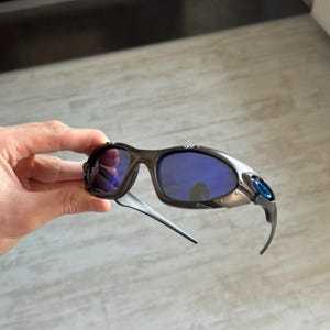 Op de afbeelding: Sportzonnebril met een zwart montuur, blauwe glazen en zilveren accenten. De zonnebril heeft een gebogen ontwerp en blauwe details op de veren. De glazen hebben een reflecterend, spiegelend uiterlijk.