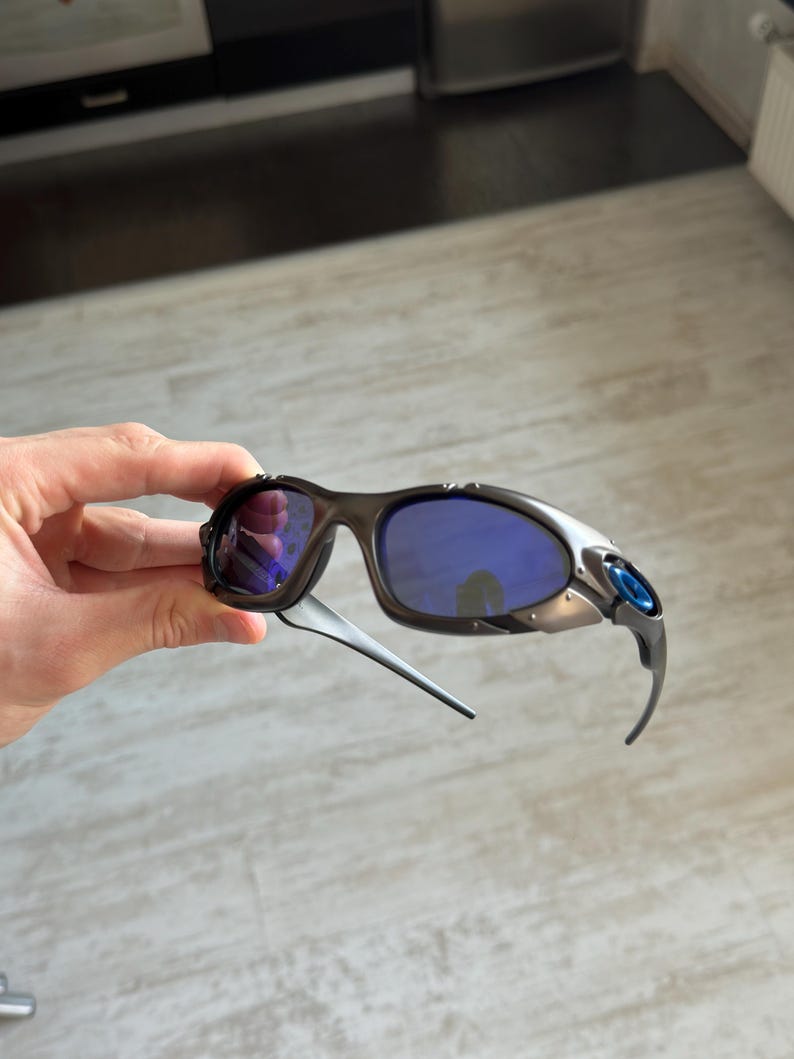 Op de afbeelding: Sportzonnebril met een zwart montuur, blauwe glazen en zilveren accenten. De zonnebril heeft een gebogen ontwerp en blauwe details op de veren. De glazen hebben een reflecterend, spiegelend uiterlijk.