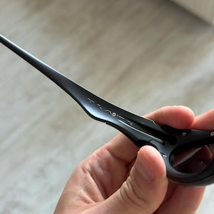 Op de afbeelding: Zwarte zonnebril met een strak, modern design. De zonnebril heeft een donker montuur en donkere glazen, met de merknaam op de poot. De zonnebril wordt in een hand gehouden.