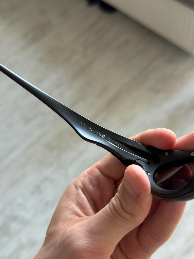 Op de afbeelding: Zwarte zonnebril met een strak, modern design. De zonnebril heeft een donker montuur en donkere glazen, met de merknaam op de poot. De zonnebril wordt in een hand gehouden.