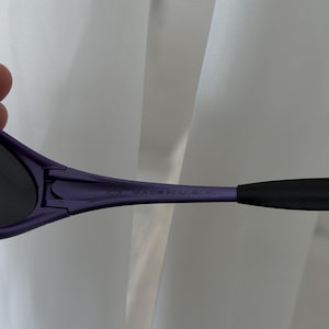 May include: Close-up of purple sunglasses with black arms. The text "CE MADE IN U.S.A." is printed on the arm of the sunglasses. The sunglasses are set against a white background.