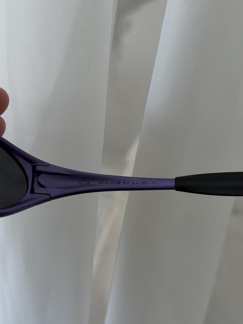 May include: Close-up of purple sunglasses with black arms. The text "CE MADE IN U.S.A." is printed on the arm of the sunglasses. The sunglasses are set against a white background.