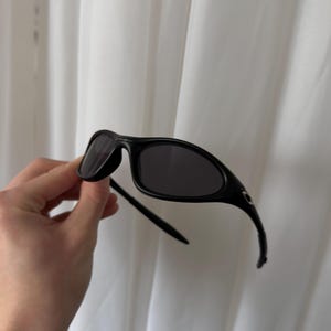 K&ouml;nnte beinhalten: Schwarze Sonnenbrille mit gebogenem Rahmen und dunklen Gl&auml;sern. Die Sonnenbrille hat ein schlankes, modernes Design. Die B&uuml;gel der Sonnenbrille kr&uuml;mmen sich zu den Ohren hin. Die Sonnenbrille wird vor einem wei&szlig;en Hintergrund gehalten.