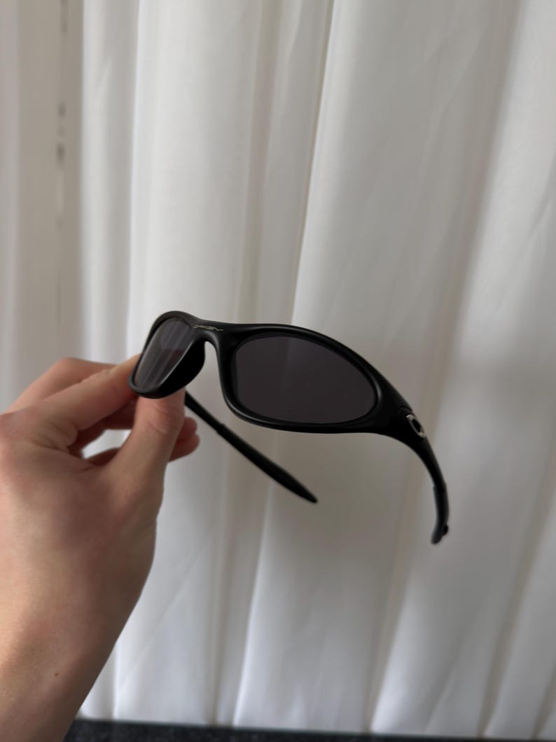 K&ouml;nnte beinhalten: Schwarze Sonnenbrille mit gebogenem Rahmen und dunklen Gl&auml;sern. Die Sonnenbrille hat ein schlankes, modernes Design. Die B&uuml;gel der Sonnenbrille kr&uuml;mmen sich zu den Ohren hin. Die Sonnenbrille wird vor einem wei&szlig;en Hintergrund gehalten.