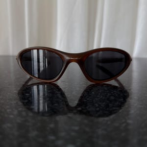 May include: Brown sunglasses with dark lenses. The sunglasses have a curved frame and a sleek, modern design. The Oakley logo is visible on the right arm of the sunglasses. The sunglasses are resting on a dark, reflective surface.