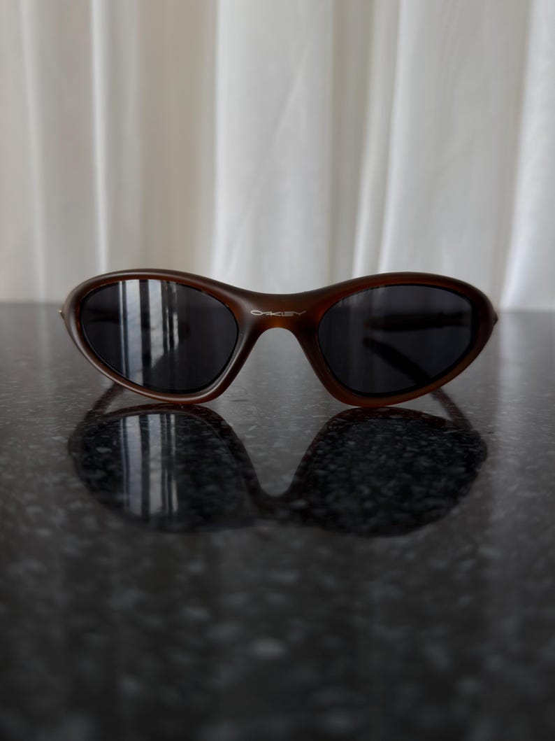 May include: Brown sunglasses with dark lenses. The sunglasses have a curved frame and a sleek, modern design. The Oakley logo is visible on the right arm of the sunglasses. The sunglasses are resting on a dark, reflective surface.