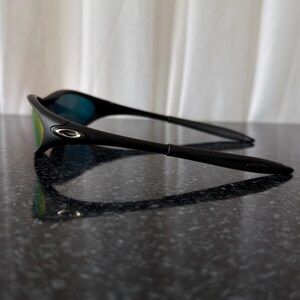 May include: Black sunglasses with a sleek, modern design. The lenses have a mirrored finish with green and blue hues. The frame is a matte black, and the arms are thin and curved. The sunglasses are resting on a dark, reflective surface.