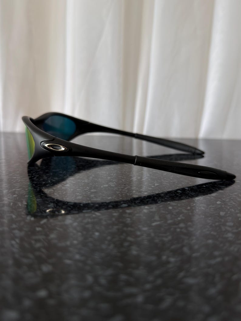 May include: Black sunglasses with a sleek, modern design. The lenses have a mirrored finish with green and blue hues. The frame is a matte black, and the arms are thin and curved. The sunglasses are resting on a dark, reflective surface.