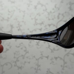 K&ouml;nnte beinhalten: Nahaufnahme einer schwarzen Sonnenbrille mit blauem Akzent am Rahmen. Die Sonnenbrille hat dunkle Gl&auml;ser und ein schlankes, modernes Design. Die B&uuml;gel sind d&uuml;nn und reichen bis zu den Ohren.
