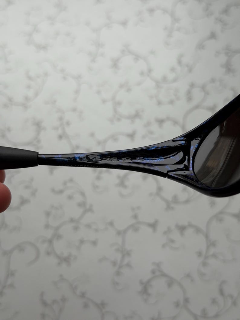 K&ouml;nnte beinhalten: Nahaufnahme einer schwarzen Sonnenbrille mit blauem Akzent am Rahmen. Die Sonnenbrille hat dunkle Gl&auml;ser und ein schlankes, modernes Design. Die B&uuml;gel sind d&uuml;nn und reichen bis zu den Ohren.