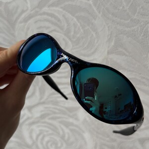 K&ouml;nnte beinhalten: Brain Dead Sonnenbrille mit blau verspiegelten Gl&auml;sern. Die Fassungen sind dunkelblau mit einem gesprenkelten Muster. Die Sonnenbrille hat eine einzigartige, abgerundete Form und ein schlankes, modernes Design.