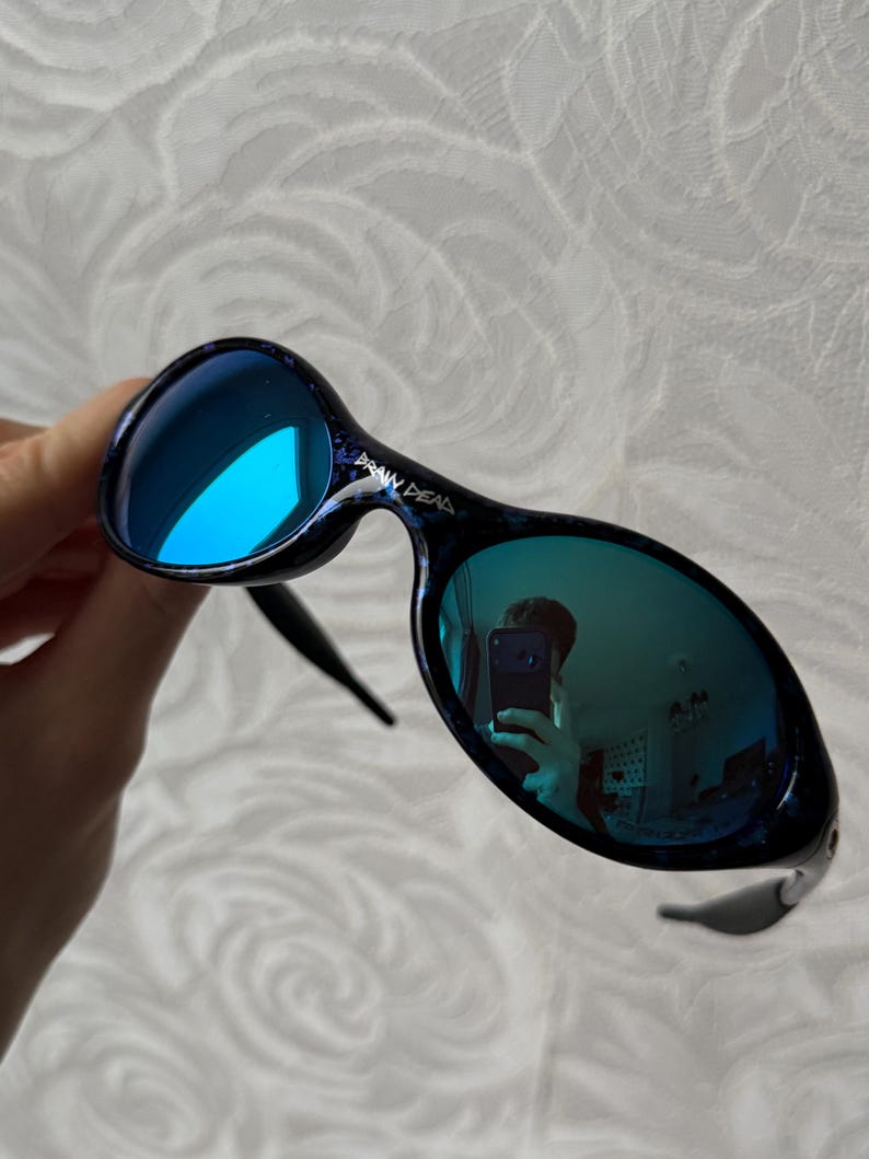 K&ouml;nnte beinhalten: Brain Dead Sonnenbrille mit blau verspiegelten Gl&auml;sern. Die Fassungen sind dunkelblau mit einem gesprenkelten Muster. Die Sonnenbrille hat eine einzigartige, abgerundete Form und ein schlankes, modernes Design.