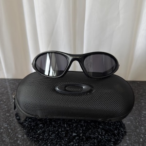 K&ouml;nnte beinhalten: Schwarze Sonnenbrille mit dunklen Gl&auml;sern liegt auf einem schwarzen, strukturierten Etui. Die Sonnenbrille hat einen geschwungenen Rahmen und ein modernes Design. Das Etui hat ein Logo und einen Rei&szlig;verschluss, was auf Schutz f&uuml;r die Brille hindeutet.