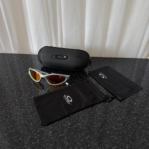Puede incluir: Gafas de sol con montura plateada y lentes naranjas, junto con un estuche negro. Las gafas incluyen una bolsa con cord&oacute;n negra y un pa&ntilde;o de limpieza, todos con el logotipo de la marca. Los art&iacute;culos est&aacute;n sobre una superficie oscura y moteada.