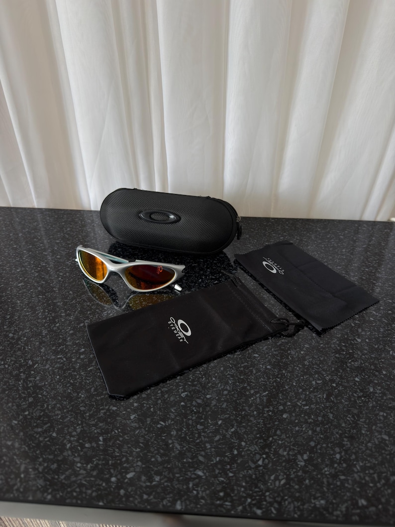 Puede incluir: Gafas de sol con montura plateada y lentes naranjas, junto con un estuche negro. Las gafas incluyen una bolsa con cord&oacute;n negra y un pa&ntilde;o de limpieza, todos con el logotipo de la marca. Los art&iacute;culos est&aacute;n sobre una superficie oscura y moteada.