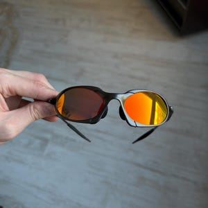 May include: Black sunglasses with orange-tinted lenses. The sunglasses have a sleek, modern design with a black frame and curved temples. The lenses reflect a bright orange color, creating a bold and stylish look.