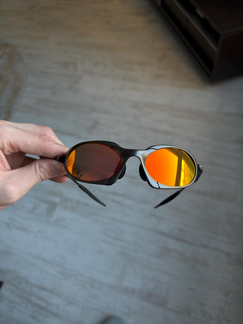 May include: Black sunglasses with orange-tinted lenses. The sunglasses have a sleek, modern design with a black frame and curved temples. The lenses reflect a bright orange color, creating a bold and stylish look.