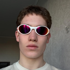May include: White oval sunglasses with red-tinted lenses. The sunglasses have a white frame with red accents on the temples. The lenses reflect a bright pink color.