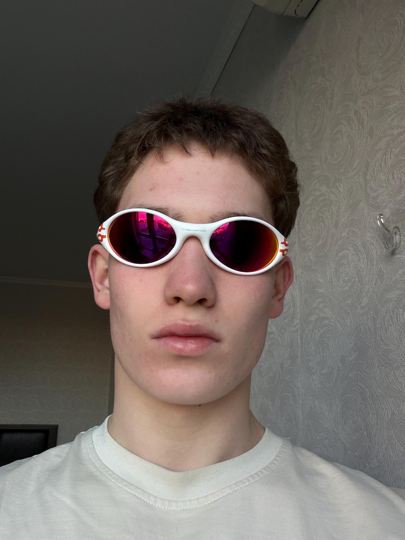 May include: White oval sunglasses with red-tinted lenses. The sunglasses have a white frame with red accents on the temples. The lenses reflect a bright pink color.