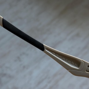 May include: Close-up of a pair of beige-colored sunglasses. The arm of the glasses is visible, with a black rubber grip and the word "straight" printed on the frame. The glasses have a modern, sporty design.