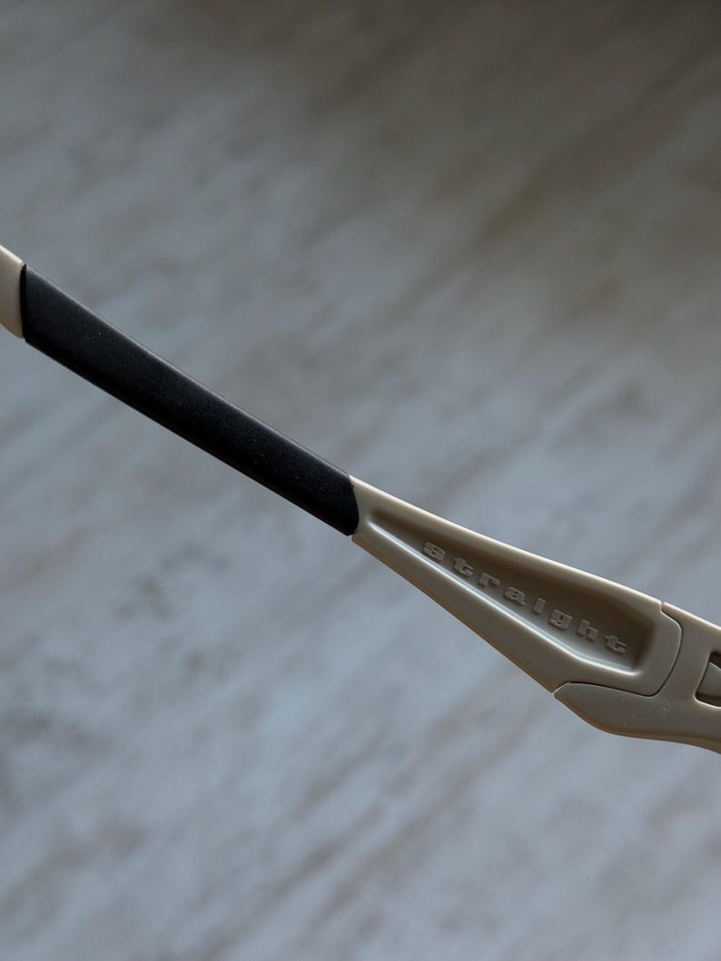 May include: Close-up of a pair of beige-colored sunglasses. The arm of the glasses is visible, with a black rubber grip and the word "straight" printed on the frame. The glasses have a modern, sporty design.