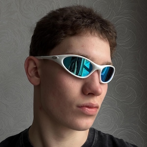 May include: White-framed sunglasses with blue reflective lenses. The sporty eyewear has a sleek, curved design. The lenses reflect a blue hue, and the frames are a bright white colour. The sunglasses are a modern accessory.
