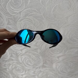 K&ouml;nnte beinhalten: Schwarze ovale Sonnenbrille mit blau verspiegelten Gl&auml;sern. Die Sonnenbrille hat ein schlankes Design mit einem dunklen Rahmen und gebogenen B&uuml;geln. Die Gl&auml;ser reflektieren einen hellblauen Farbton und erzeugen einen auff&auml;lligen Kontrast.