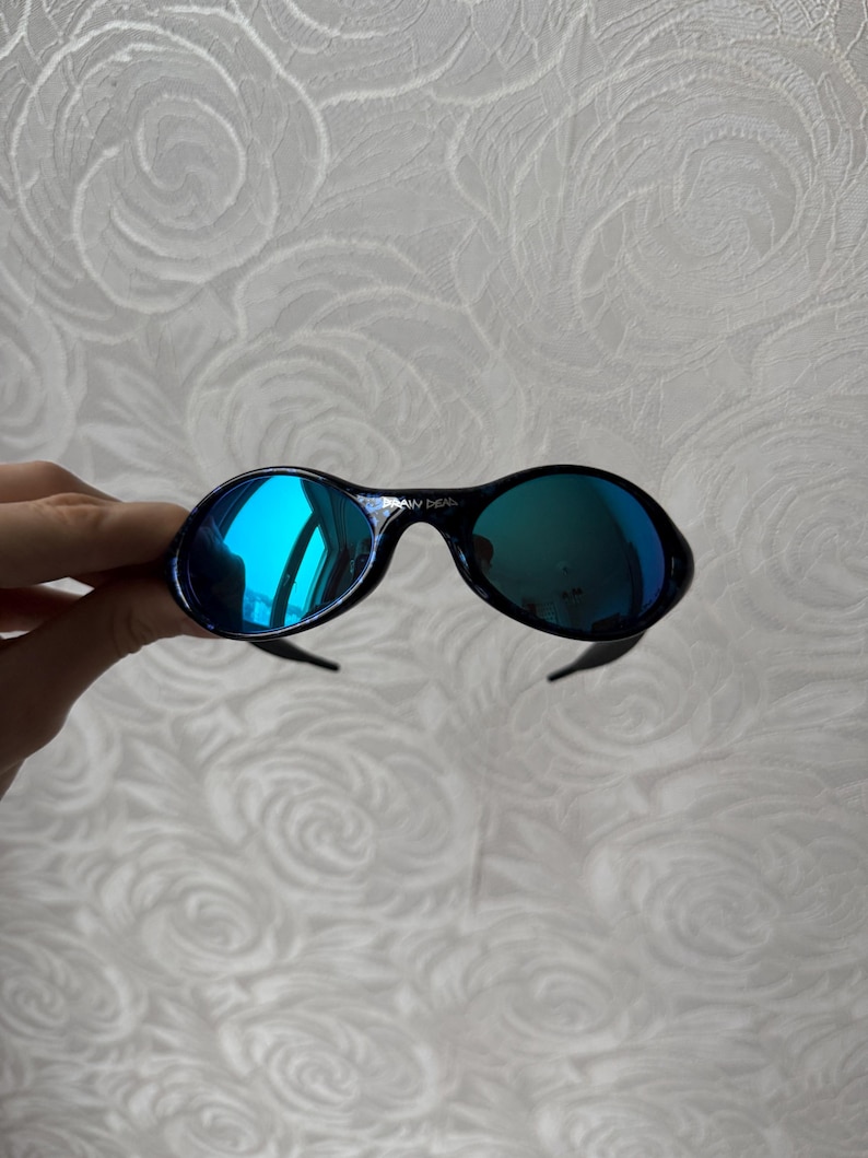 K&ouml;nnte beinhalten: Schwarze ovale Sonnenbrille mit blau verspiegelten Gl&auml;sern. Die Sonnenbrille hat ein schlankes Design mit einem dunklen Rahmen und gebogenen B&uuml;geln. Die Gl&auml;ser reflektieren einen hellblauen Farbton und erzeugen einen auff&auml;lligen Kontrast.