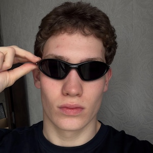 K&ouml;nnte beinhalten: Schwarze ovale Sonnenbrille mit dunklen Gl&auml;sern. Die Sonnenbrille hat ein schlankes, modernes Design. Der Rahmen ist gl&auml;nzend schwarz. Die Sonnenbrille wird leicht angehoben.