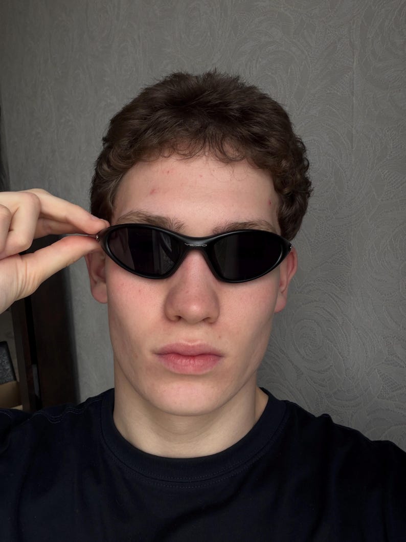 K&ouml;nnte beinhalten: Schwarze ovale Sonnenbrille mit dunklen Gl&auml;sern. Die Sonnenbrille hat ein schlankes, modernes Design. Der Rahmen ist gl&auml;nzend schwarz. Die Sonnenbrille wird leicht angehoben.