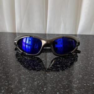 Op de afbeelding: Een zilverkleurige zonnebril met blauwe glazen. De zonnebril heeft een sportief, omsluitend ontwerp. De glazen reflecteren de omgeving. De zonnebril ligt op een donker, gespikkeld oppervlak.
