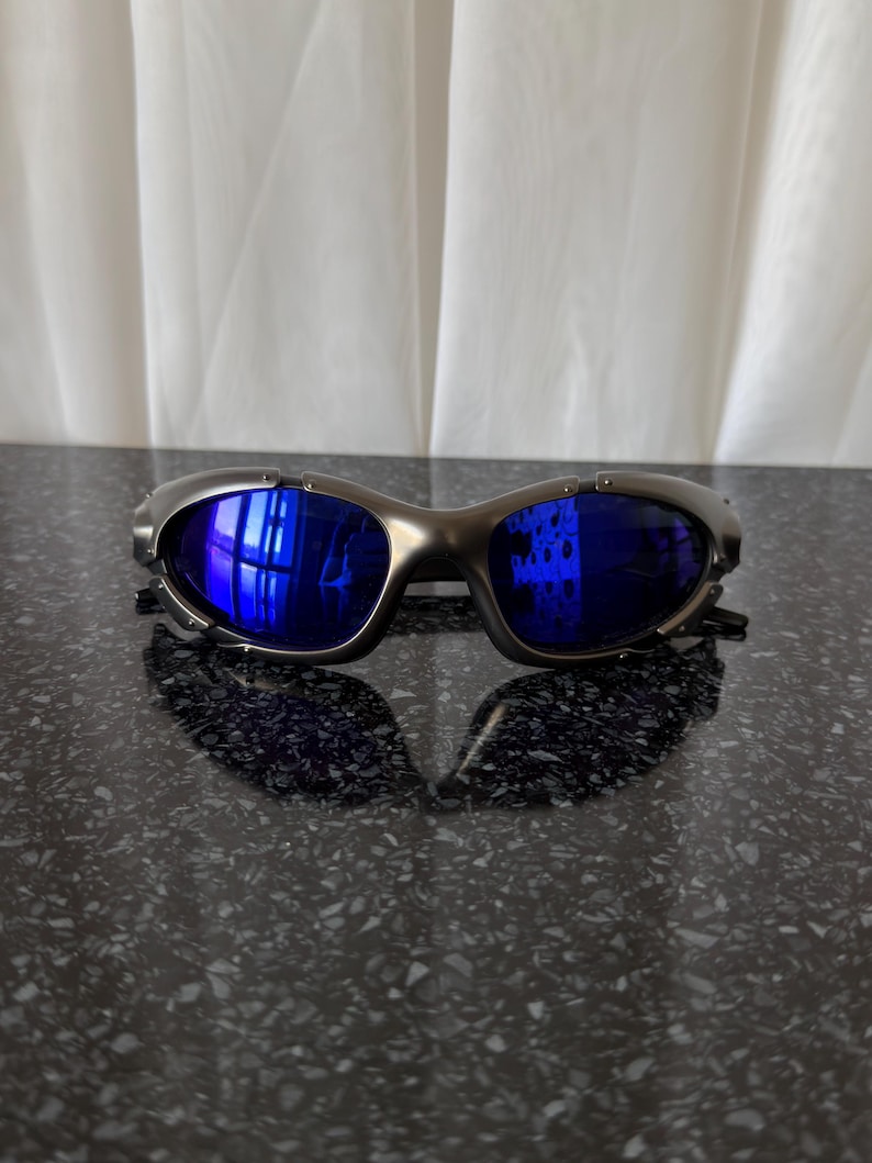 Op de afbeelding: Een zilverkleurige zonnebril met blauwe glazen. De zonnebril heeft een sportief, omsluitend ontwerp. De glazen reflecteren de omgeving. De zonnebril ligt op een donker, gespikkeld oppervlak.