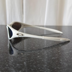 Puede incluir: Gafas de sol blancas con un dise&ntilde;o elegante. Presentan una montura blanca, lentes oscuras y un logotipo plateado. Las patillas son largas y delgadas. El aspecto general es deportivo y moderno, ideal para actividades al aire libre.