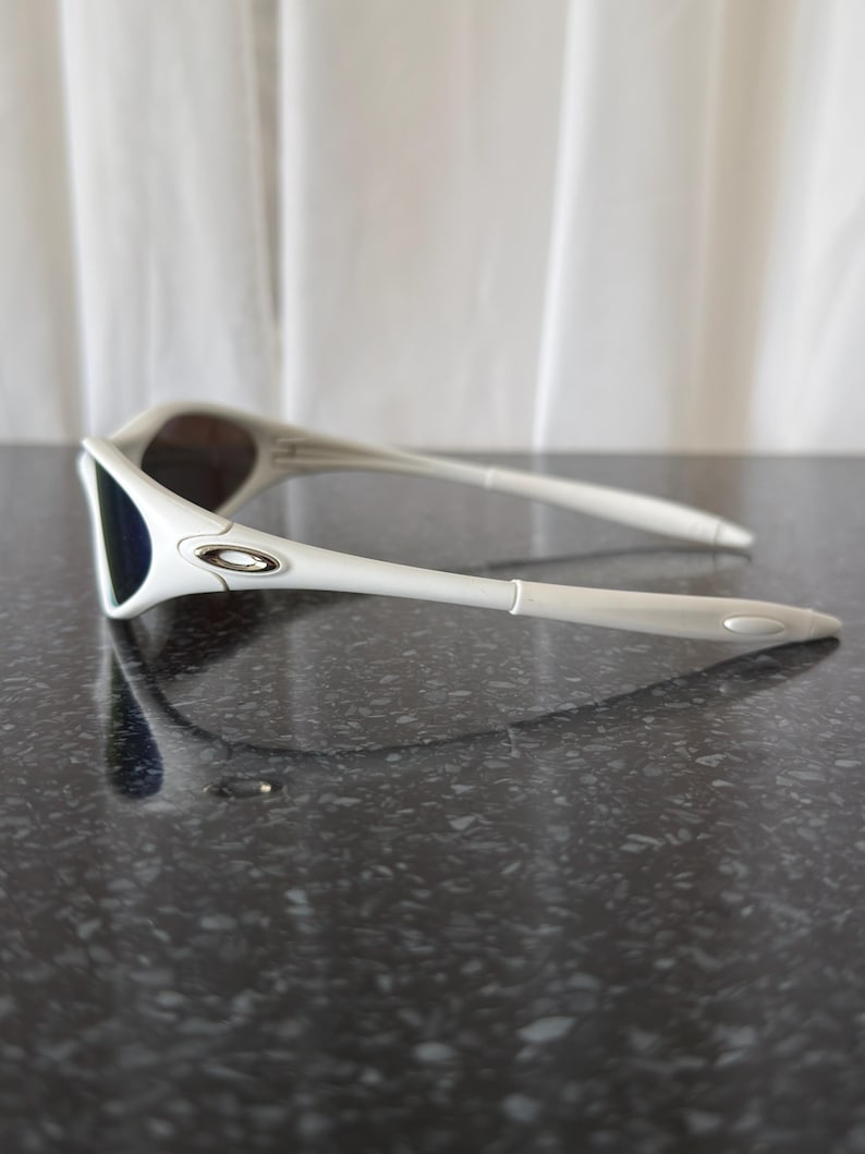Puede incluir: Gafas de sol blancas con un dise&ntilde;o elegante. Presentan una montura blanca, lentes oscuras y un logotipo plateado. Las patillas son largas y delgadas. El aspecto general es deportivo y moderno, ideal para actividades al aire libre.