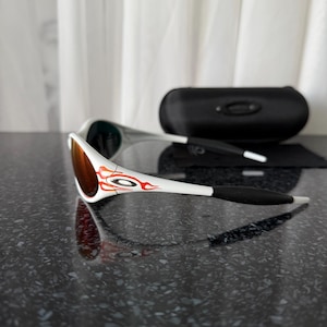 May include: White and black sunglasses with a red and orange design on the arms. The lenses are a reddish-brown color. A black sunglasses case and a black cleaning cloth are also visible.