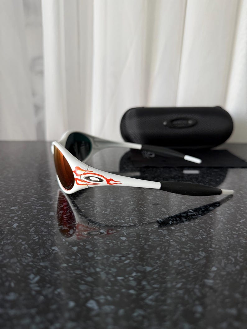 May include: White and black sunglasses with a red and orange design on the arms. The lenses are a reddish-brown color. A black sunglasses case and a black cleaning cloth are also visible.