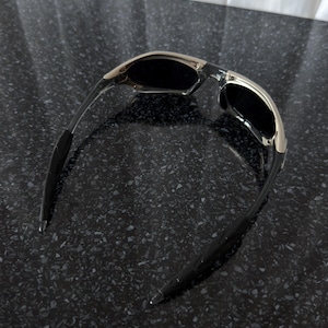 May include: Sunglasses with a silver frame and black lenses. The temples are black and curve inward. The sunglasses are set against a dark, speckled surface.