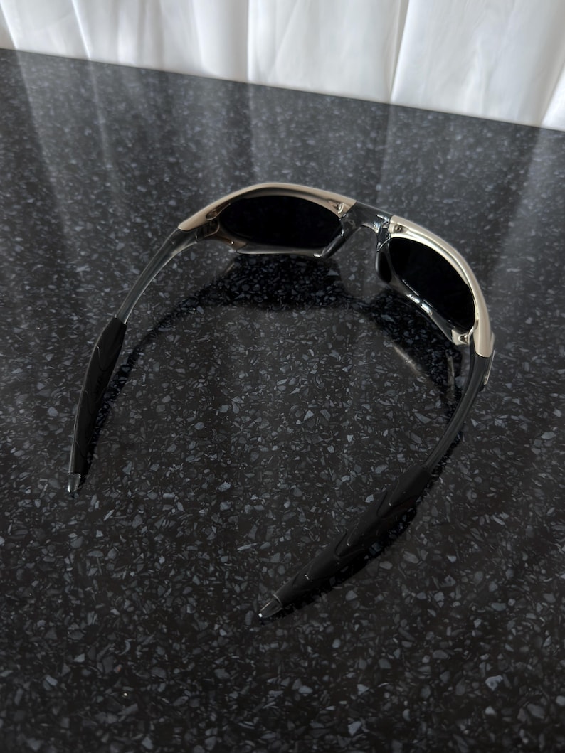 May include: Sunglasses with a silver frame and black lenses. The temples are black and curve inward. The sunglasses are set against a dark, speckled surface.