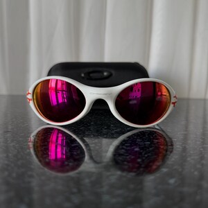May include: White sunglasses with red mirrored lenses and a black case. The sunglasses have a white frame with red accents on the temples. The lenses reflect a pinkish hue. The case is rectangular and made of a matte black material.