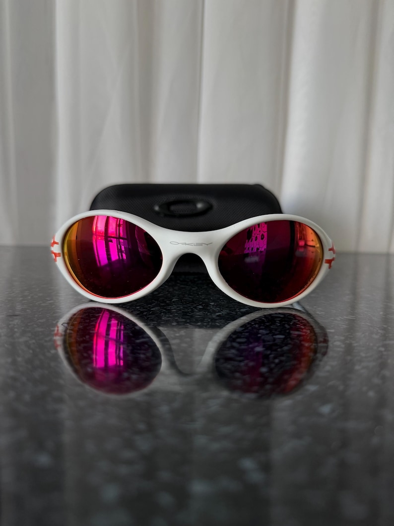 May include: White sunglasses with red mirrored lenses and a black case. The sunglasses have a white frame with red accents on the temples. The lenses reflect a pinkish hue. The case is rectangular and made of a matte black material.