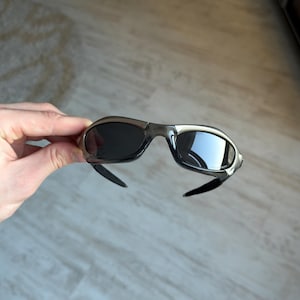 May include: A pair of sporty sunglasses with a silver frame and dark lenses. The sunglasses have a curved design and black temples. The lenses reflect the surrounding environment.
