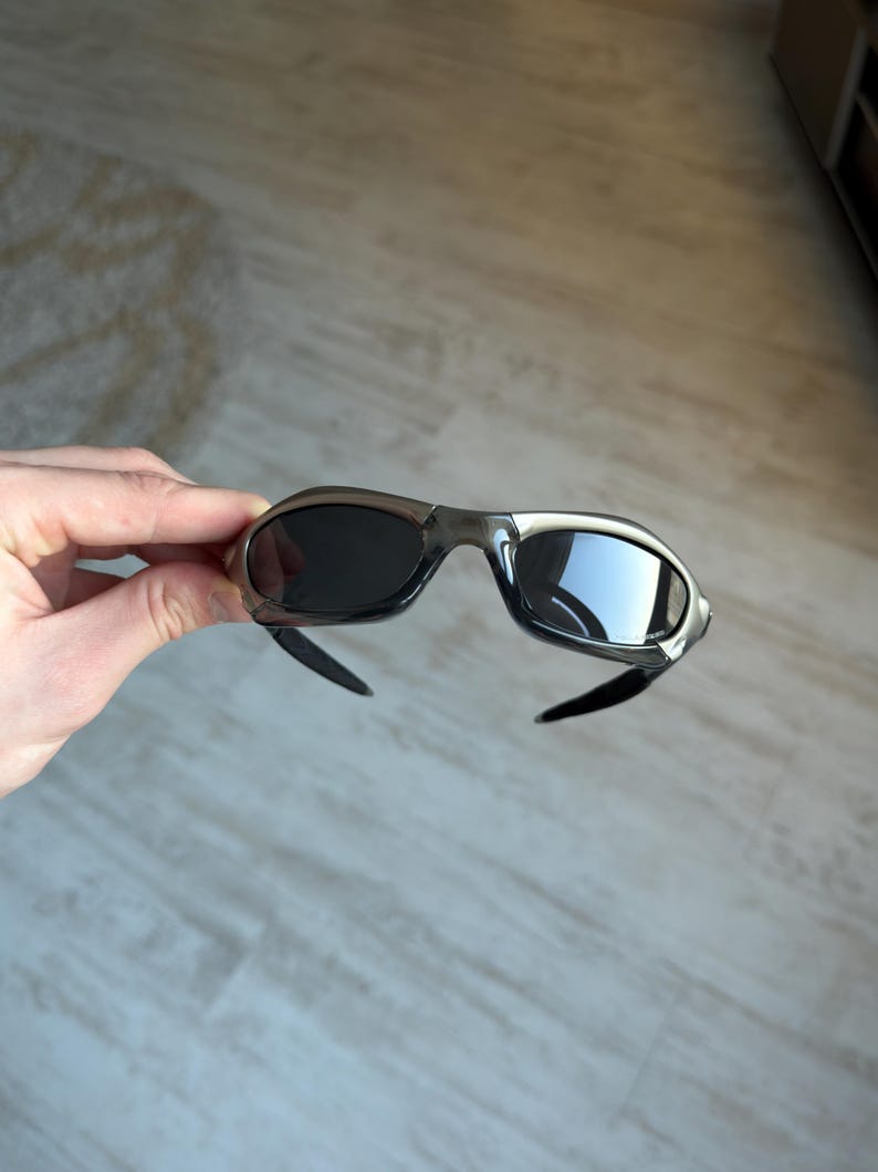 May include: A pair of sporty sunglasses with a silver frame and dark lenses. The sunglasses have a curved design and black temples. The lenses reflect the surrounding environment.
