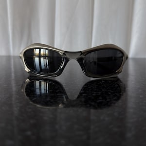 May include: Sleek, metallic-framed sunglasses with dark lenses. The sunglasses have a curved design and reflect the surrounding environment. The lenses are dark, providing a contrast to the metallic frame. The sunglasses are resting on a reflective surface.