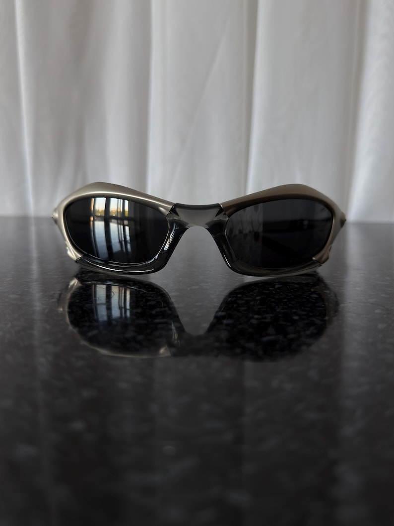 May include: Sleek, metallic-framed sunglasses with dark lenses. The sunglasses have a curved design and reflect the surrounding environment. The lenses are dark, providing a contrast to the metallic frame. The sunglasses are resting on a reflective surface.